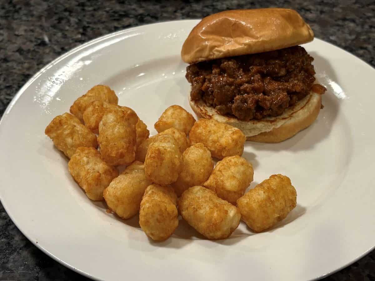 A plate with a classic Sloppy Joes sandwich on a bun and a serving of golden tater tots, placed on a speckled countertop.