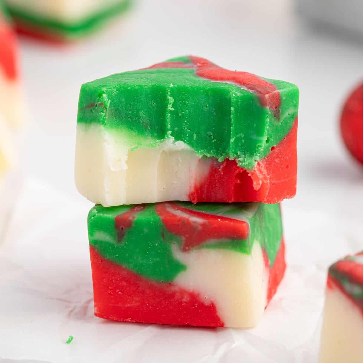Two pieces of red, green, and white swirled Christmas Fudge are stacked on top of each other, with a bite taken from the top piece, sitting on white parchment paper.