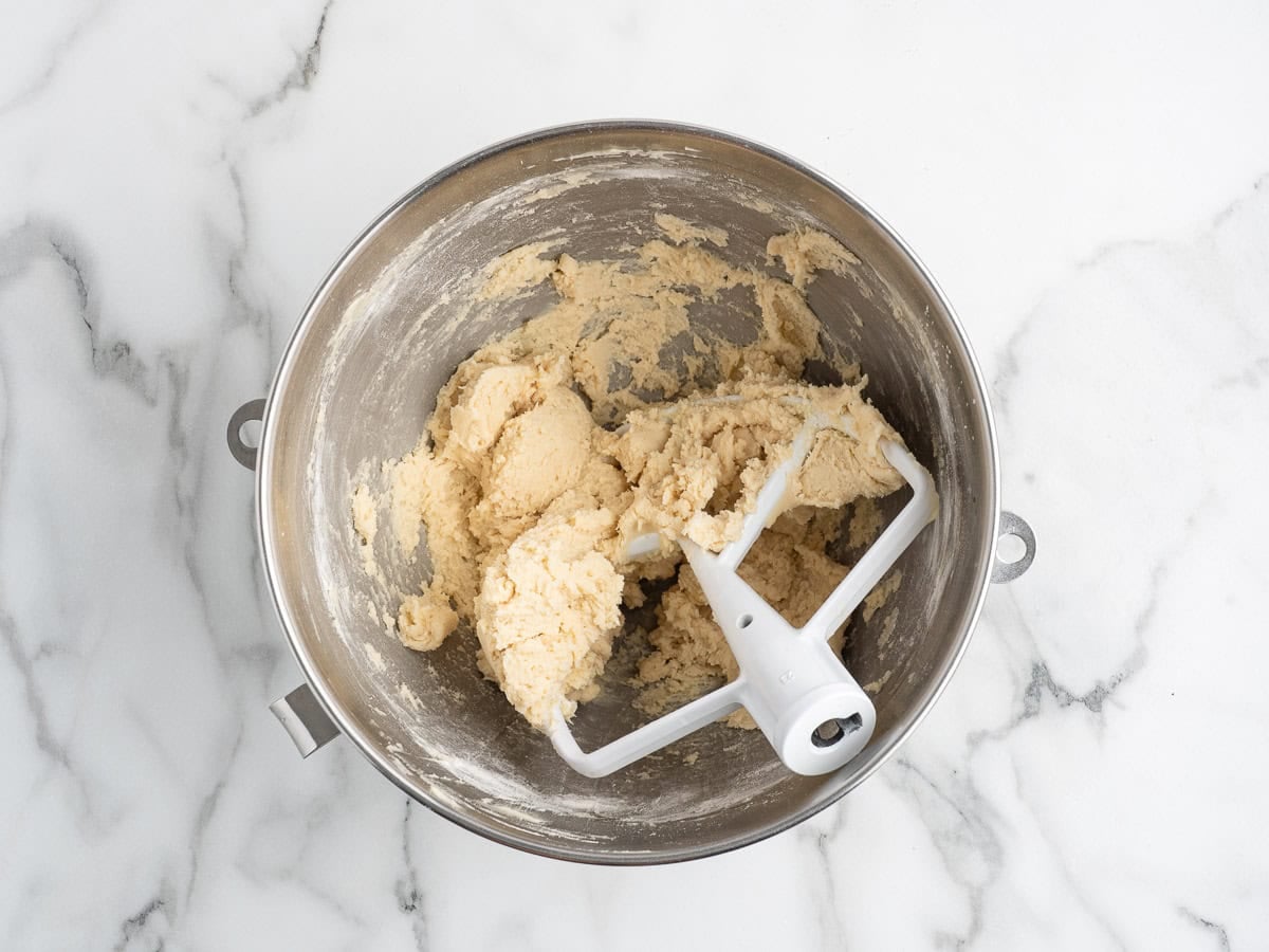 A metal mixing bowl with pale Christmas Shortbread Cookies dough being mixed by a white paddle attachment, sitting on a white marble surface.