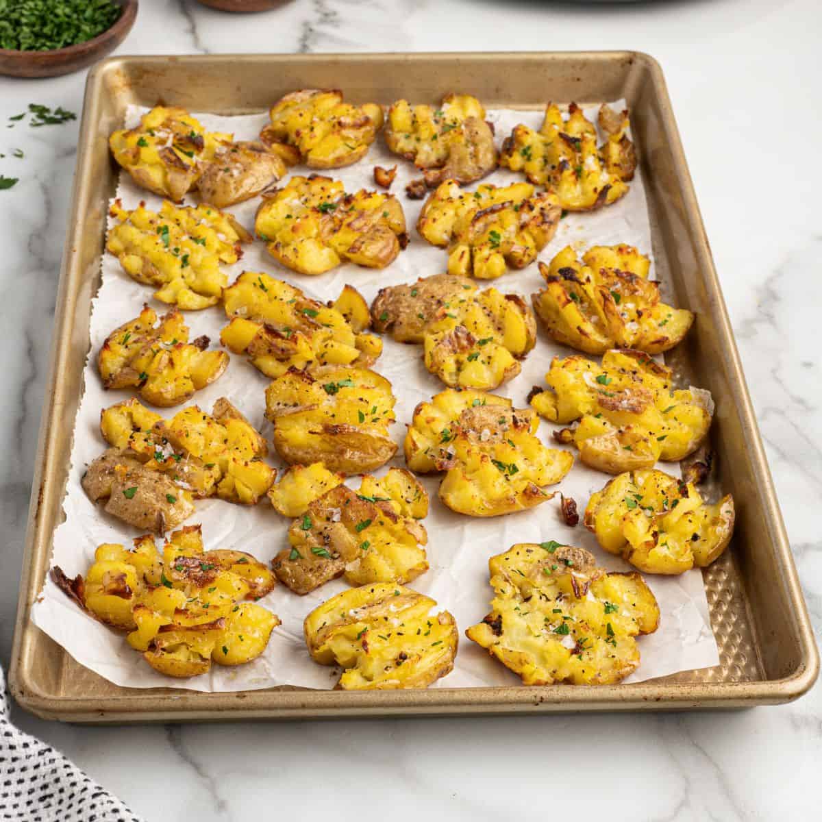 Golden crispy smashed potatoes sprinkled with herbs are arranged on a parchment-lined baking sheet, ready to serve.