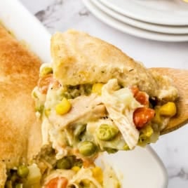 A wooden spoon lifts a serving of creamy chicken pot pie with mixed vegetables and a golden, flaky crust from a white baking dish. Plates and utensils sit in the background.